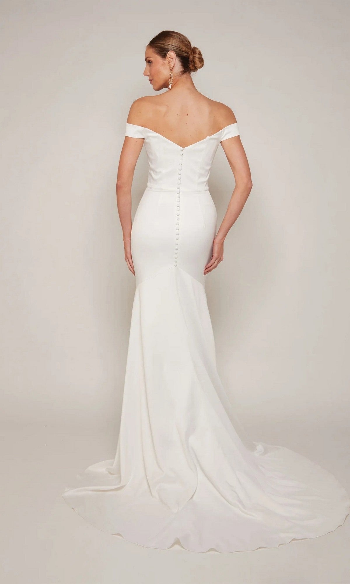 A woman faces away, wearing the Alyce 7098 long formal dress—an off-the-shoulder white gown with back button details and a long train. Her hair is styled in a sleek low bun.